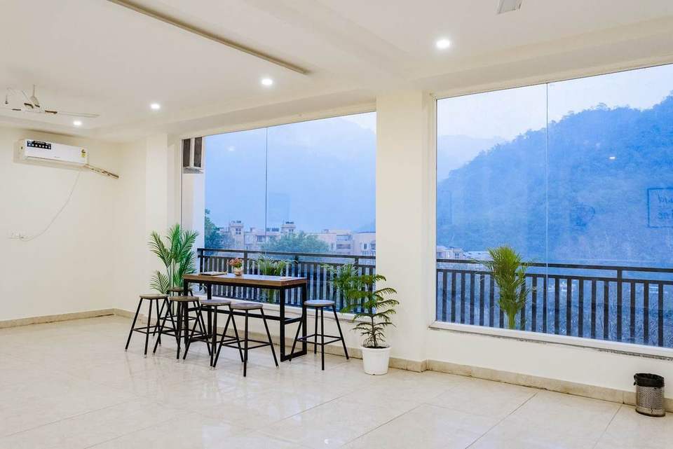 Sunlit lounge with glass walls, plants and a high table looking at the hills at Alpine Retreat, Rishikesh